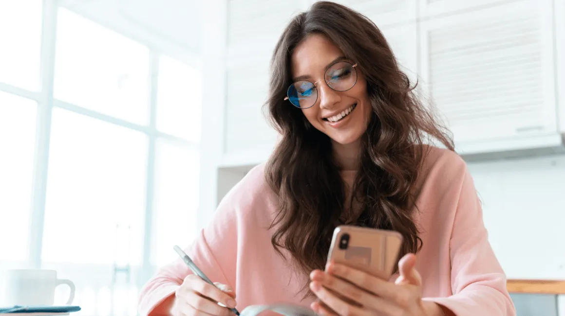 Kobieta w okularach uśmiecha się, robi notatki i trzyma telefon w ręku
